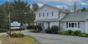 A gray sided with green shutters office building surrounded by evergreens and a pavement parking lot in front.