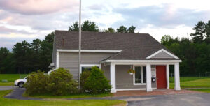A gray and white sided small office building with flag pole in front and hanging plants.