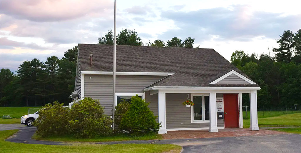 A gray and white sided small office building with flag pole in front and hanging plants.